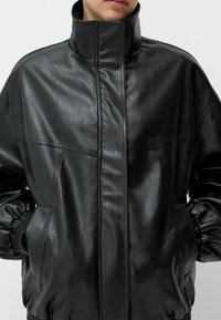 Jachetă de piele neagră cu guler înalt, având o textură fină, manșete elastice și buzunare frontale. Design minimalist cu linii curate.