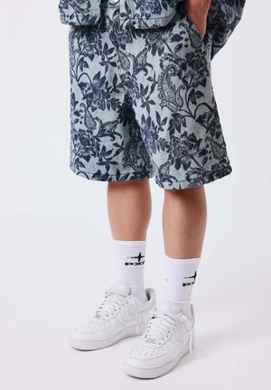Beine, die graue Shorts mit Blumenmuster, weiße Kniestrümpfe mit schwarzem Text und weiße Nike-Sneaker mit gebundenen Schnürsenkeln tragen.