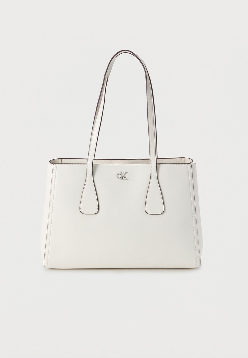 Λευκή τσάντα tote από συνθετικό δέρμα με διπλές λαβές, αντίθετη σκουρόχρωμη επένδυση και ασημί λογότυπο στη μπροστινή πλευρά. Ομαλή υφή, ορθογώνιο σχήμα.