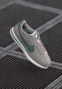 Nike kedas ar gaiši pelēku augšdaļu, zaļu akcentu uz Swoosh un bēša teksturēta zamša purngala. Redzamas kurpes auklas un retro zoles dizains.