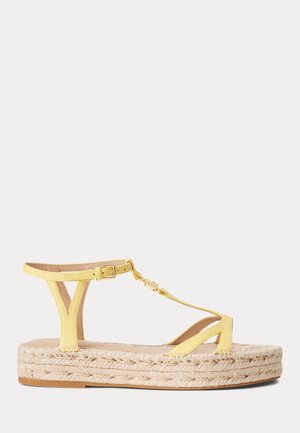 Gul läder T-rem plattformssandal med flätad jutesula och guldfärgade spänne på ankelrem, sidovy på vit bakgrund.