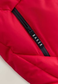 Chaqueta acolchada roja con cremallera negra y una etiqueta con el texto "BAKER". La tela parece suave con un brillo sutil.