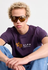 Violet trumpas rankovėmis marškinėliai su balta ir auksine "ALPHA INDUSTRY" logo, derinami su rudos spalvos akinių nuo saulės ir šviesiai mėlynomis džinsinėmis kelnėmis.