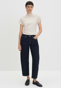 Haut côtelé beige clair à manches courtes associé à un pantalon en denim foncé ample avec cinq poches et fermeture éclair. Chaussures noires à enfiler.