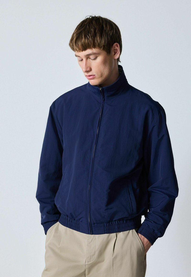 Jeune homme aux cheveux courts et bruns portant une veste bleu marine zippée et un pantalon beige, les mains dans les poches, regardant vers le bas.