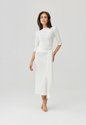 Žena v bílé midi šatech s dlouhým rukávem a zavazováním na boku, obutá v béžových sandálech na vysokém podpatku se stahovacími pásky, stojící před jednobarevným pozadím.