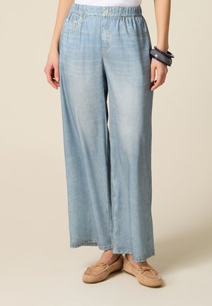 Pantalon en jean bleu clair à jambes larges avec taille élastique, poches avant, porté avec des mocassins beiges et des bracelets foncés à un poignet.