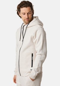 Felpa con cappuccio di un beige chiaro, con zip frontale, lacci neri, polsini a coste e tasche laterali; realizzata in un tessuto testurizzato e morbido.