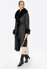 Černý semišový dlouhý kabát s velkým černým kožíškem na límci a na manžetách, zavázaný v pase, doplněný černými kozačkami po kolena a bílou kabelkou.