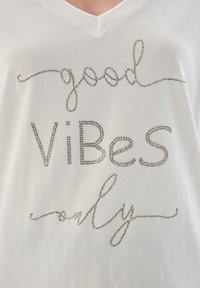 Bílý tričko s krátkým rukávem, které nese nápis "jen dobré vibrace" vyrobený ze stříbrných kamínků, má výstřih do V a hladkou strukturu.