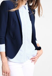 Mujer vestida con un blazer azul marino sobre una camisa de botones a rayas azul claro y blanca, con pantalones blancos, mano resting sobre la cadera.