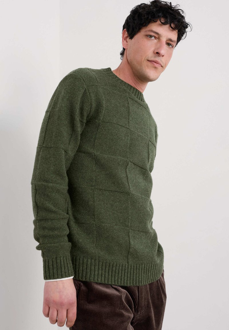 Maglione verde con texture e motivo a quadri, colletto rotondo e orlo a costine. Il modello lo indossa con pantaloni in cotone a coste marroni.