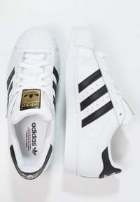 Vita lädersneakers med gummiskal tå, med tre svarta ränder och en guldfärgad logga på tungan. Texturerade sulor och slät yta.