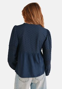 Marineblaue Bluse aus strukturiertem Stoff, mit gepufften langen Ärmeln und einem peplumartigen Bund, der ein dekoratives Diamantmuster auf der Rückseite aufweist.