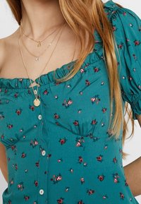 Tyrkysová květinová blůza s off-shoulder volánkovým výstřihem, krátkými nafouknutými rukávy a předními knoflíky. Materiál je lehký s hladkou texturou.