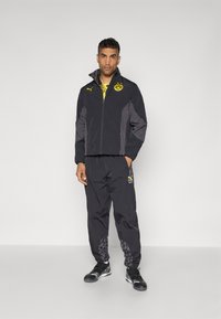 Schwarze und graue Reißverschlussjacke mit hohem Kragen, gelbem Logo und Akzenten; passende schwarze Hose mit gemusterten Unterschenkeln; schwarze Sportschuhe.