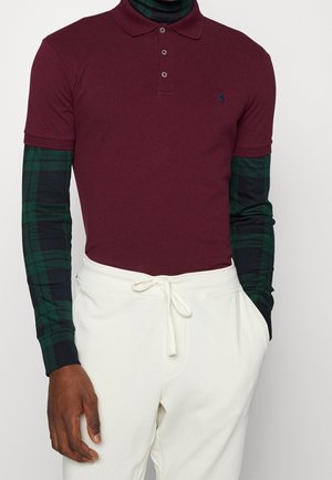 Bordeauxrotes Poloshirt mit klassischem Kragen und drei Knöpfen, darüber ein grün-schwarz kariertes Langarmhemd; weiße Hose.
