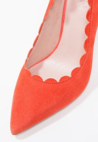 Chaussure plate en daim corail à bout pointu avec un détail de bord festonné le long de l'ouverture et une doublure intérieure lisse rose clair.