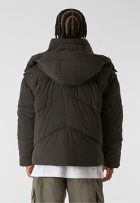 Schwarze Puffjacke mit Kapuze, mit gestepptem Muster und Zickzack-Nähten, getragen über einem weißen Hemd und Cargo-Hosen.