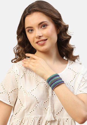 Multistrenget armbånd med blå, grønne og sølvfarvede perler, med en struktureret overflade og frynse-dekorationer. Båret på en models håndled.