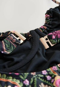 Svart simdräkt med blommönstrade detaljer har texturerade volangkanter och guldfärgade spännen. Slät tygkomposition.