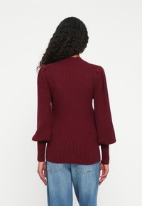 Kvinne med langt, krøllete hår iført en ribbet, burgunder genser med puffede ermer og blå jeans, stående med ryggen til på en enkel hvit bakgrunn.