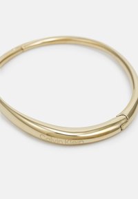 Bracciale in metallo dorato con superficie liscia e lucida e design minimalista. Presenta un logo inciso in modo discreto lungo il bordo.