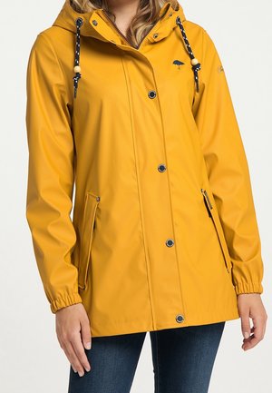 Femme portant un imperméable jaune avec des boutons-pression noirs, une capuche avec cordons, des poches zippées et à boutons, et des poignets élastiques.