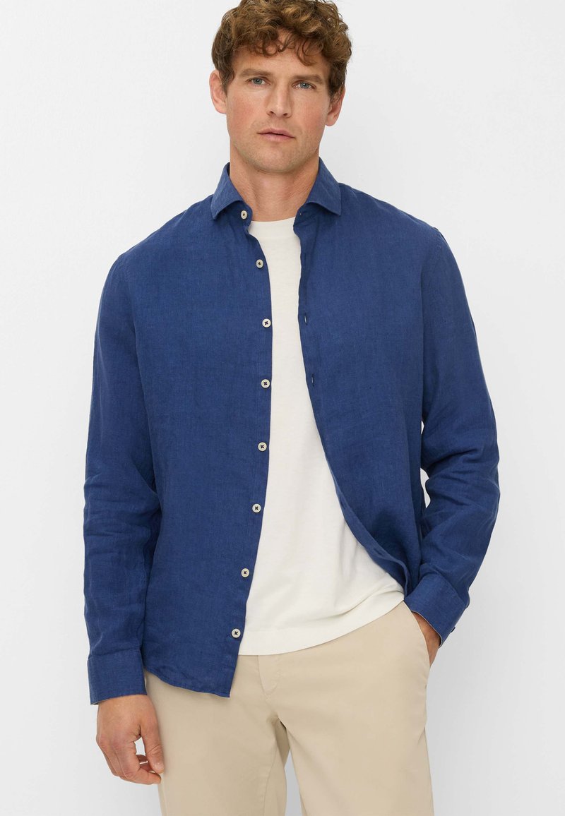 Navy blue linen shirt with a button-up front, long sleeves, and a relaxed fit, layered over a white T-shirt and paired with beige pants.