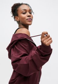 Sweat à capuche zippé bordeaux avec une coupe décontractée, doté d'un cordon de serrage, de manches larges et d'un col audacieux, sur un fond neutre.