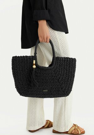 Osoba držící velkou černou tkanou kabelku s třásněmi a korálky, oblečená v texturovaných bílých kalhotách, černém topu a hnědých sandálech.