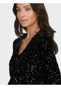Czarny top z cekinami, z dekoltem w kształcie V i długimi rękawami, o teksturowanej powierzchni z nachodzącymi na siebie błyszczącymi okrągłymi cekinami.