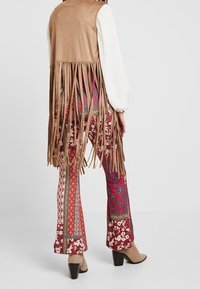 Brauner Wildlederwesten mit langen Fransen, über gemusterten Hosen in Rot und Blau mit floralen und geometrischen Designs. Das Model trägt beiges Absatzschuhe.