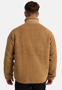 Chaqueta de forro polar texturizado marrón con un cuello alto, diseño acanalado y un ajuste relajado. La superficie parece suave y esponjosa.