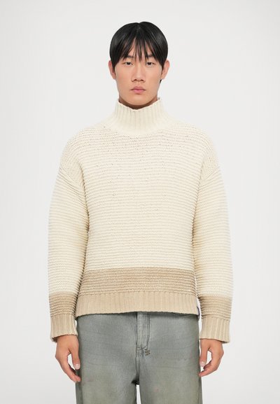 Pull en maille côtelée crème et beige avec col roulé, présentant un motif rayé texturé et une coupe ample.