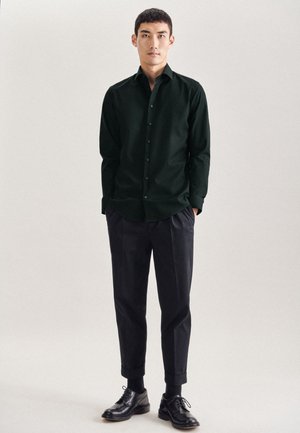 Camicetta verde scuro con bottoni e maniche lunghe, abbinata a pantaloni neri a sigaretta e scarpe nere con lacci. Design minimalista, senza patterns.