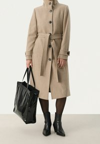 Cappotto in lana beige con colletto alto, vita con cintura e bottoni neri. Una borsa a spalla nera lucida e stivaletti neri con tacco completano il look.