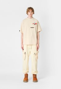 Camiseta oversize beige con detalle gráfico y logo en el pecho, acompañada de pantalones de chándal beige con bolsillos cargo con cremallera; botas marrones.