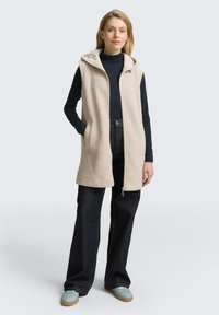 Beige Fleece-Weste mit Kapuze, Reißverschluss und Seitentaschen. Kombiniert mit dunklen Jeans und blauen Slippern. Angenehme Textur.