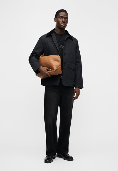 Μαύρο σακάκι με γιακά, μαύρο μπλουζάκι με λογότυπο, καφέ δερμάτινη τσάντα, σκουρόχρωμο τζιν παντελόνι, μαύρα παπούτσια. Το μοντέλο στέκεται μπροστά σε μονόχρωμο φόντο.