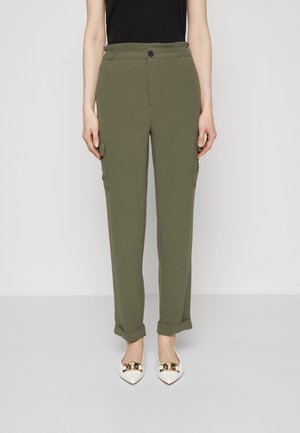 Kvinde iført olivengrønne cargobukser med opbuk ved manchetterne, sort ærmeløs top og hvide spidse flade sko med guldkæde-detaljer.