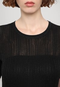 Femme portant un haut en maille côtelée noire semi-transparente avec un col rond, montré du cou à la partie supérieure de la poitrine.