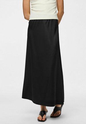 Žena v dlouhé černé sukni, bílém tílku a černých sandálech na podpatku s otevřenou špičkou stojí před jednoduchým pozadím.