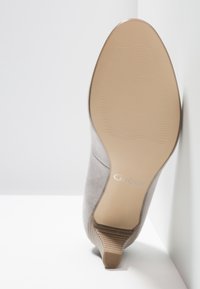 Semelle beige d'une chaussure à talon en daim gris clair appuyée contre un coin blanc avec le nom de la marque Gabor sur la semelle.
