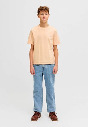 Nastoletni chłopiec stoi przodem, ubrany w brzoskwiniową koszulkę, jasnoniebieskie dżinsy oraz brązowe buty na sznurowadła na jednolitym tle.