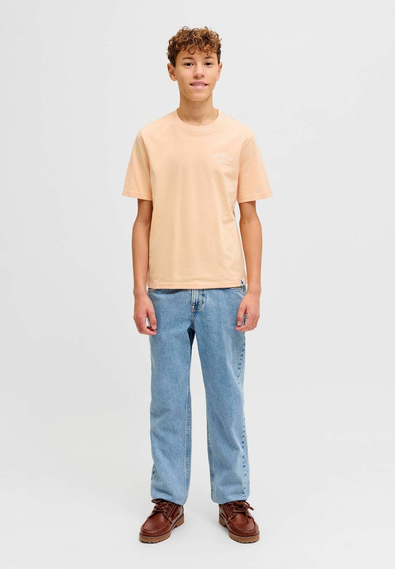 Adolescent garçon debout, faisant face à l'avant, portant un t-shirt pêche, un jean bleu clair et des chaussures marron à lacets sur un fond uni.