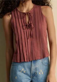 Rosa ärmelloses Oberteil mit vertikalen Falten und einer Schleife am Kragen. Kombiniert mit blauen Denim-Shorts, die mit Blumenapplikationen verziert sind.