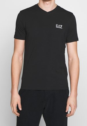 Mężczyzna ubrany w czarną koszulkę z krótkim rękawem i dekoltem w serek z białym logo EA7 Emporio Armani na lewej piersi oraz czarne spodnie.