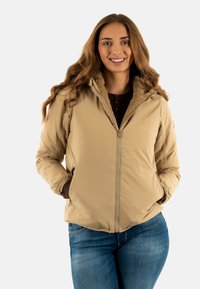 Donna che sorride con lunghi capelli, indossa una giacca beige con zip e foderata in pelliccia e jeans blu, con le mani in tasca, in piedi contro uno sfondo bianco.