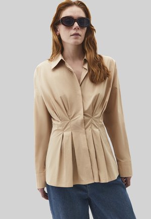 Kvinde med langt rødt hår iført skildpaddeskal-solbriller, beige plisseret skjorte og blå jeans foran en ensfarvet grå baggrund.